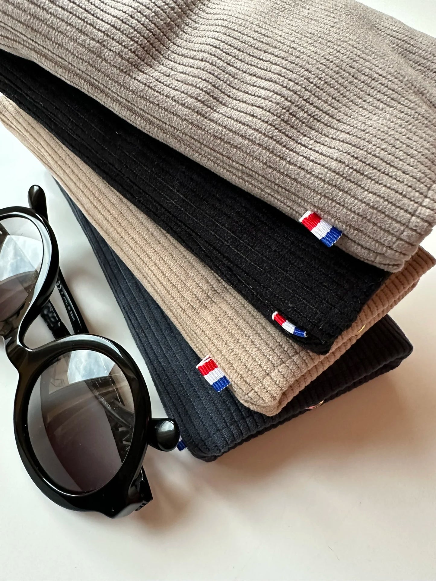 L'étui à lunettes en velours cotelé taupe, posé sur le noir, le beige et le bleu canard.