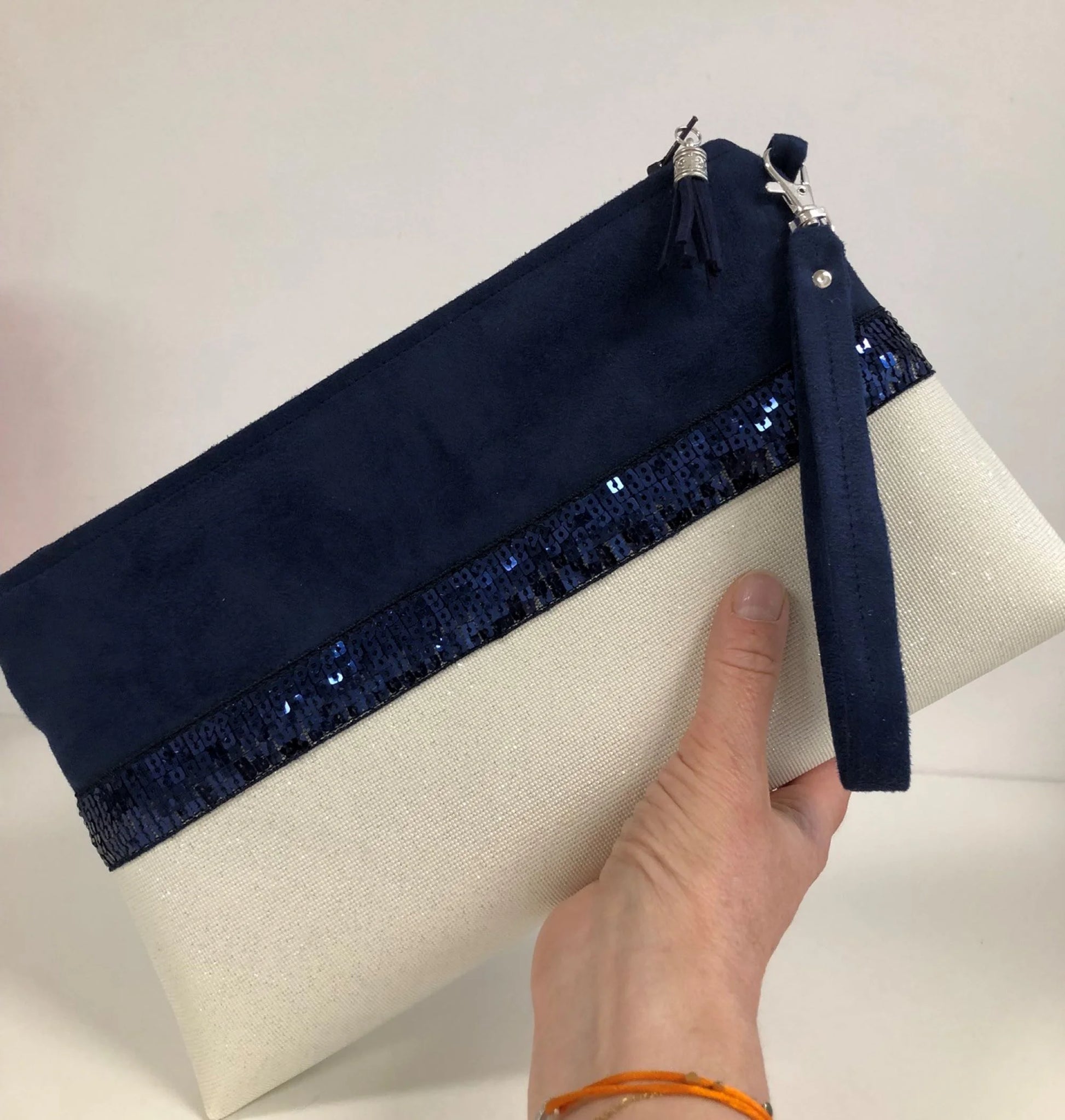 La pochette Isa zippée bleu marine et blanc à paillettes avec sa dragonne amovible, dans la main.