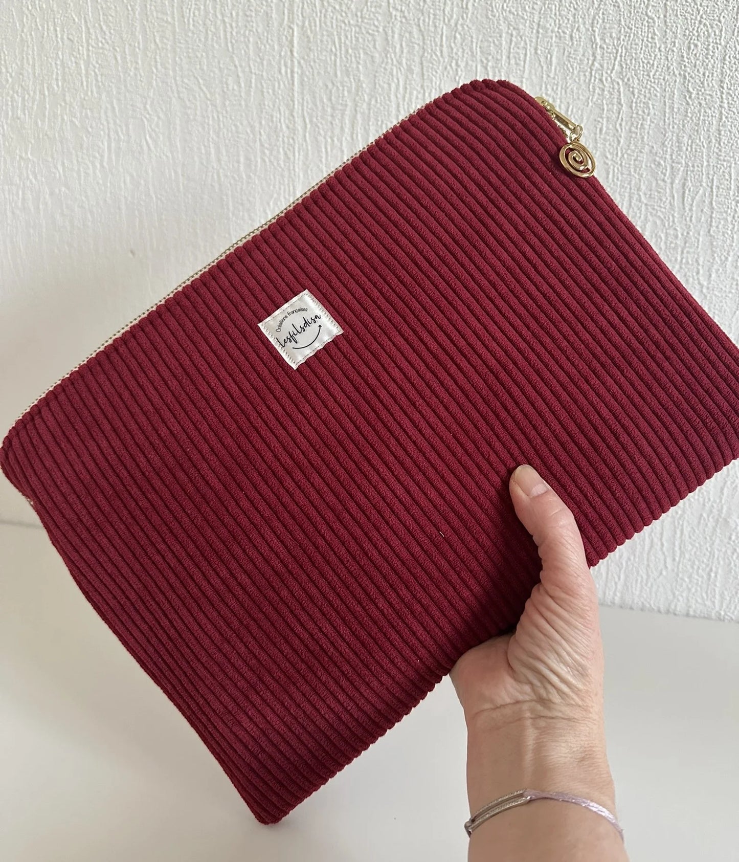 La pochette ordinateur en velours cotelé bordeaux, format 13 pouces, tenue à la main.