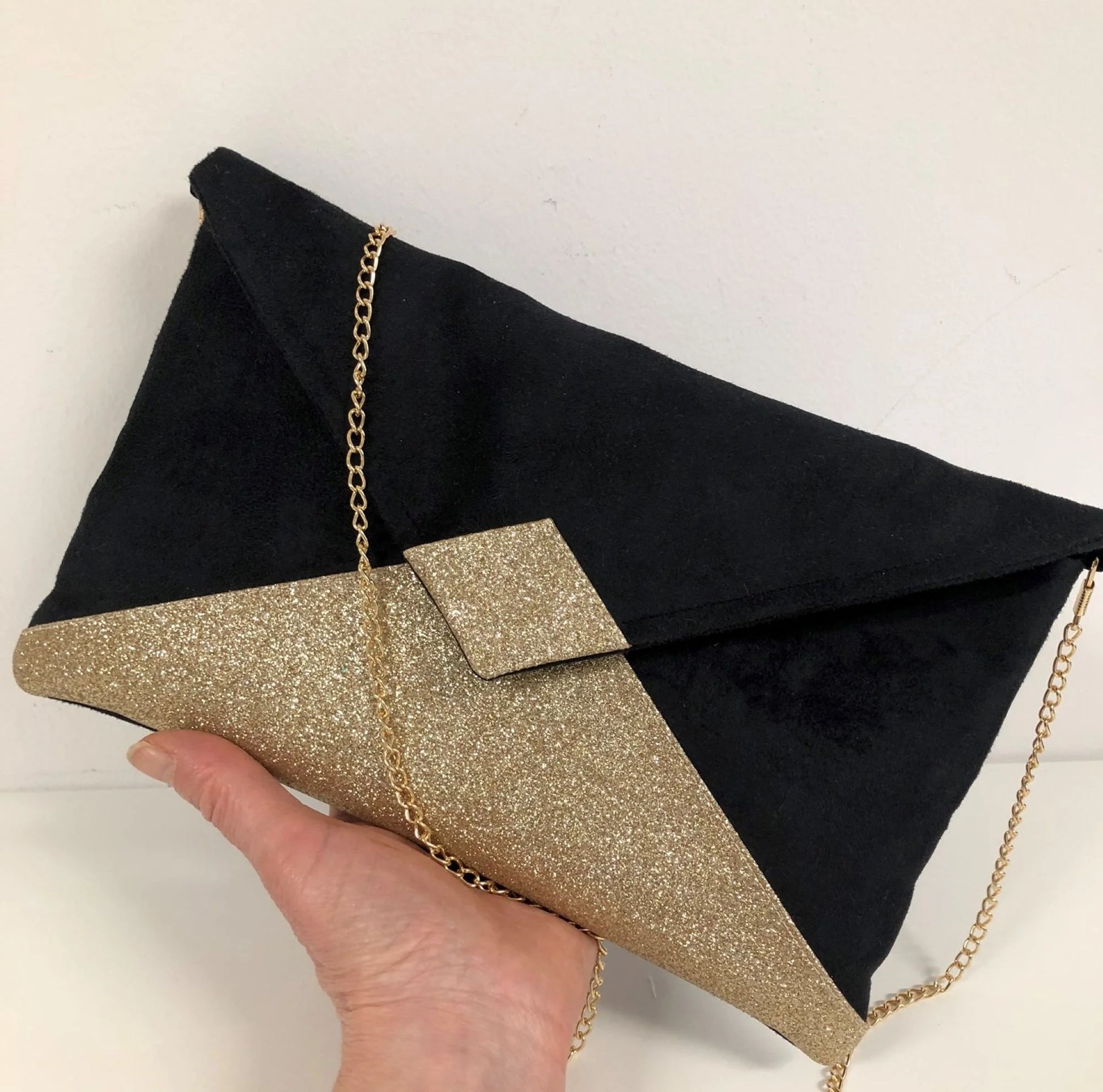Le sac pochette Isa noir à paillettes dorées, avec chainette amovible, tenu en main.
