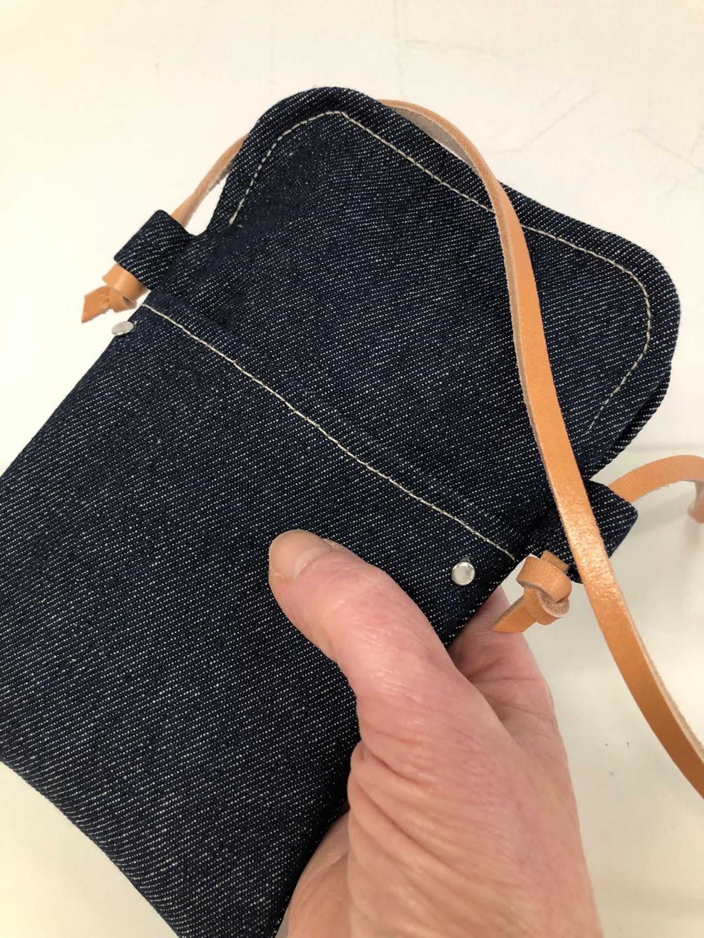 La  pochette bandoulière téléphone en denim et cuir naturel, tenue à la main.