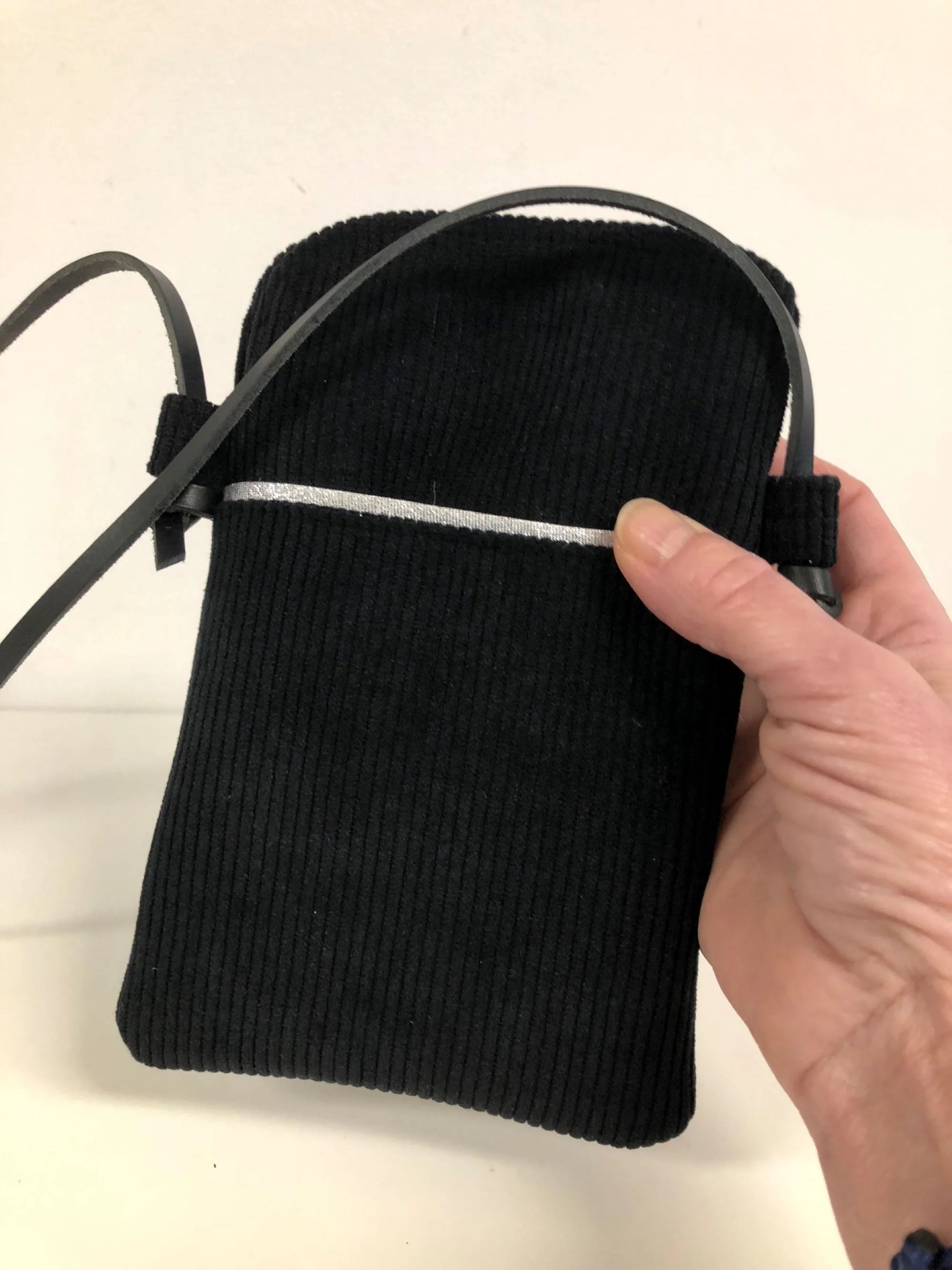 La pochette téléphone bandoulière en velours cotelé noir et cuir, tenue en main.