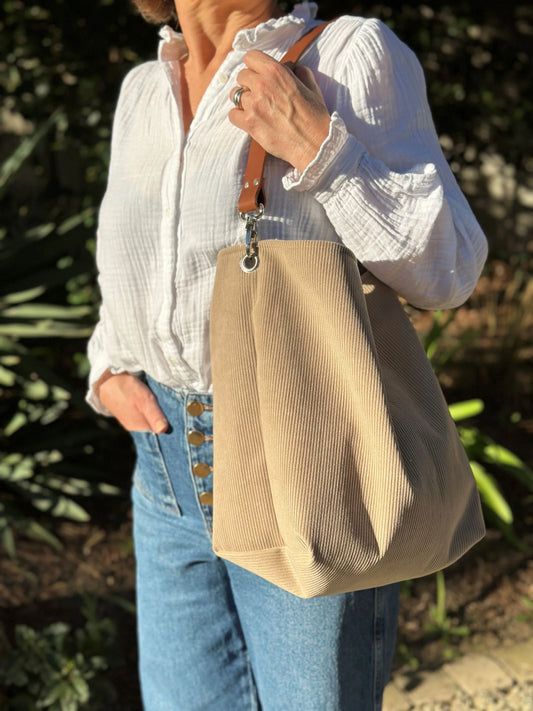 Le sac hobo en velours cotelé beige et son anse en cuir marron, porté à l'épaule.