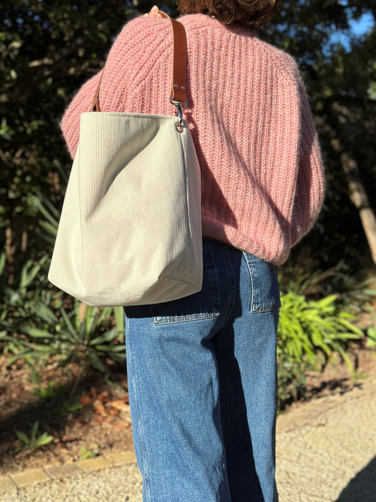 Le sac hobo en velours cotelé ivoire et son anse en cuir fauve, porté à l'épaule.