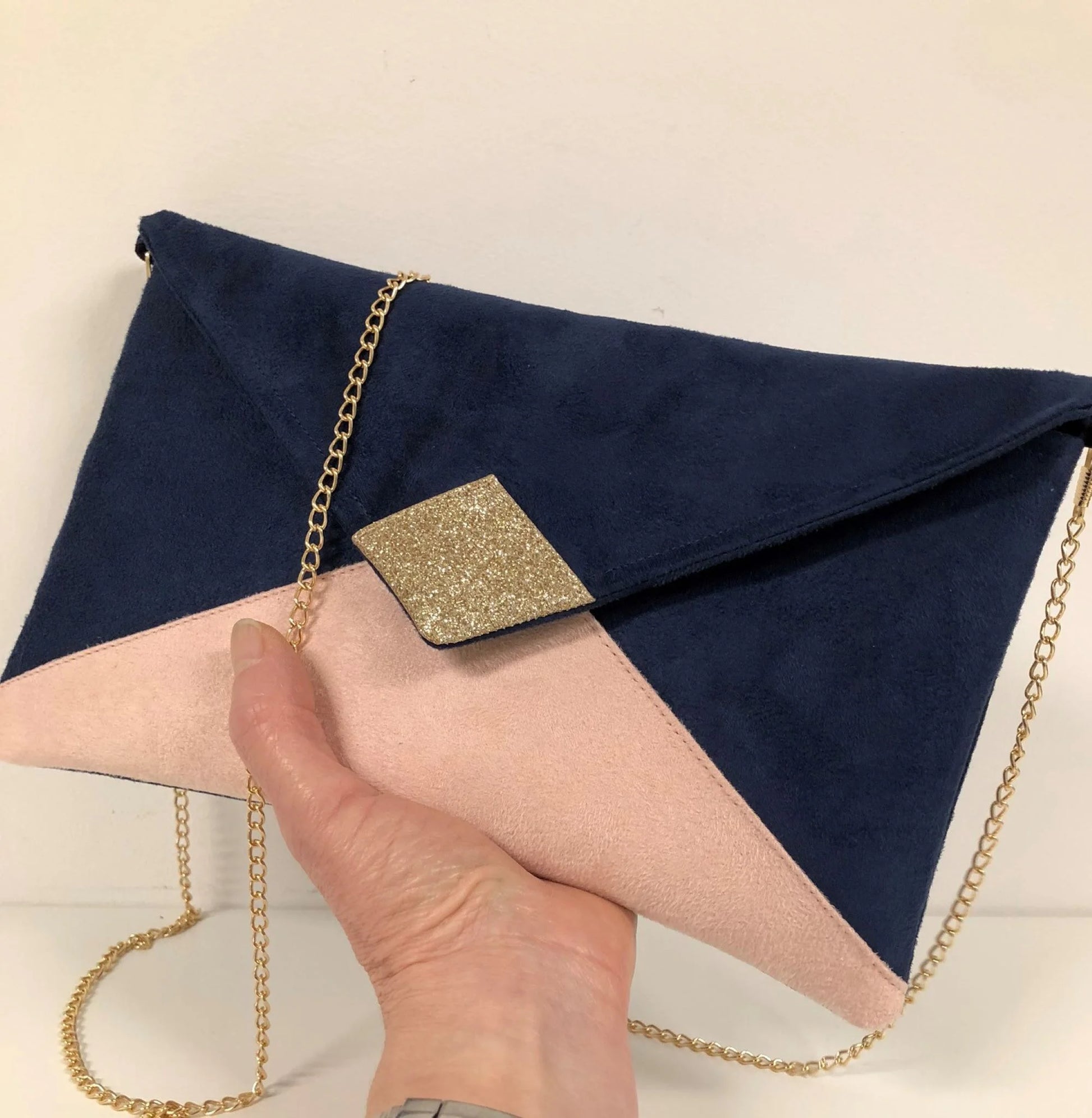Le sac pochette Isa bleu marine et rose poudré à paillettes dorées, avec chainette amovible, tenu en main.